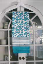 Load image into Gallery viewer, Teal and white patterned tablecloth hanging on a white hanger against a glass door.